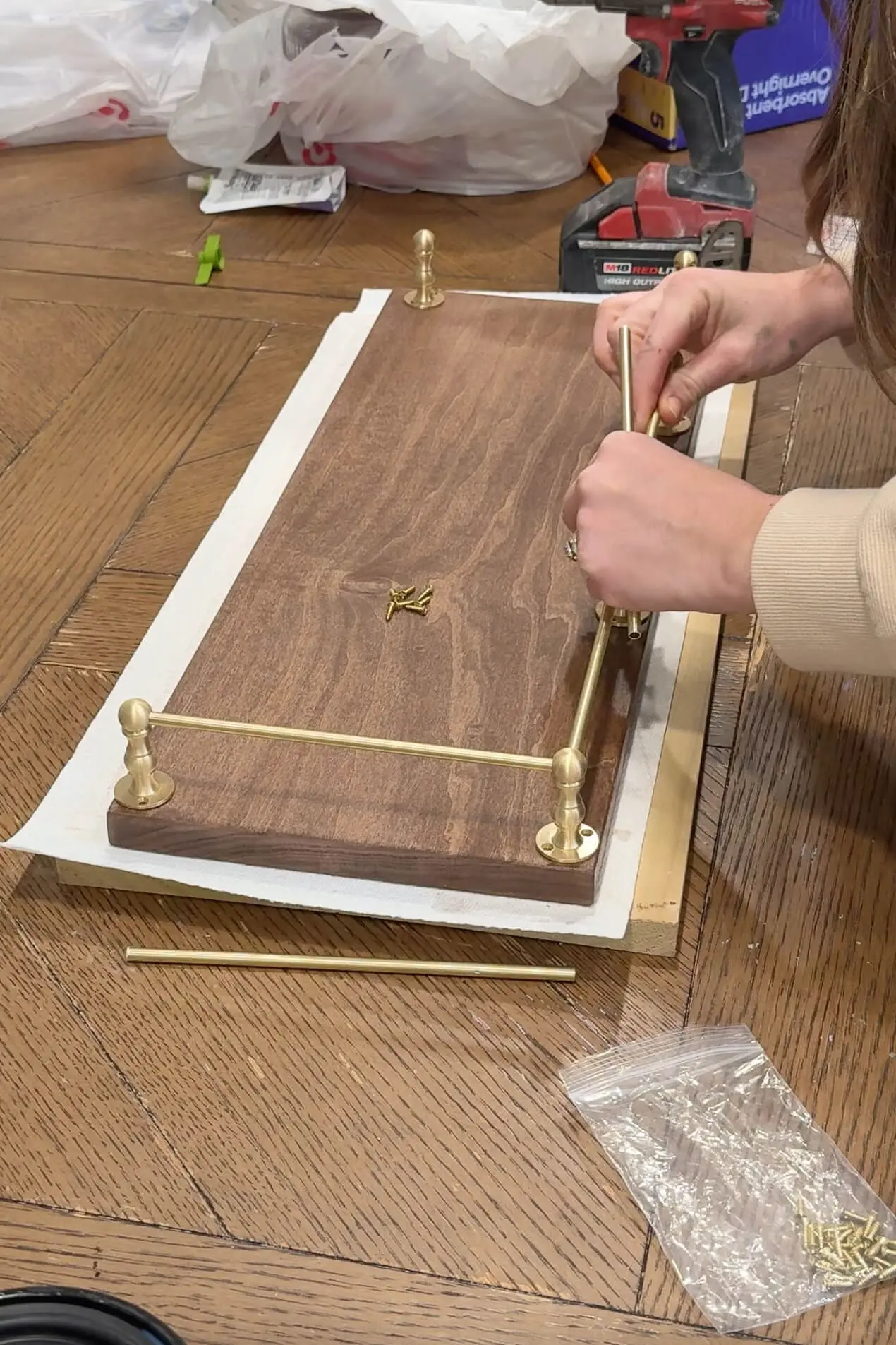 A partially completed wooden shelf, now stained and sealed, with brass rods being attached for decorative detail. Small screws and tools are visible.